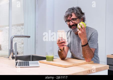 Happy senior man with apple using smart phone at kitchen island Stock Photo