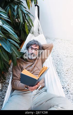 Man reading book by lying on hammock in backyard Stock Photo