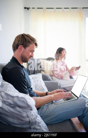Freelancer working on laptop with woman using mobile phone at home Stock Photo
