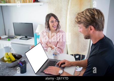 Happy young freelancer gesturing discussing with woman using laptop sitting at dining table Stock Photo