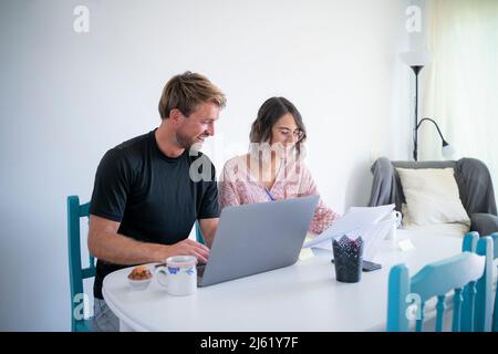 Smiling freelancer with laptop sitting by woman reading documents sitting at dining table Stock Photo