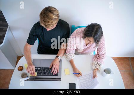 Young freelancer writing on document sitting by man using laptop at dining table Stock Photo