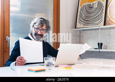 Smiling freelancer reading documents sitting with laptop at table Stock Photo