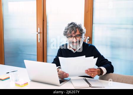 Happy freelancer reading documents sitting with laptop at home Stock Photo