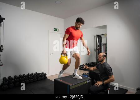 Coach guiding young man exercising with kettlebell at health club Stock Photo