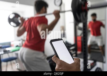 Hand of coach holding smart phone near young man lifting barbell at gym Stock Photo