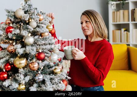 Woman with coffee cup looking at ornament on Christmas tree Stock Photo