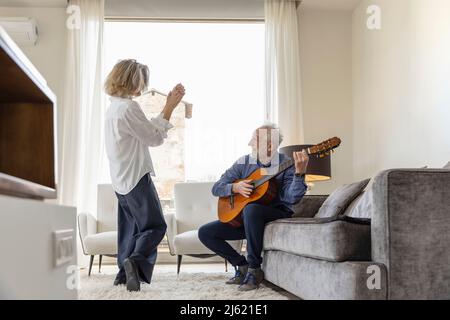 Senior man playing guitar looking at woman dancing in living room at home Stock Photo