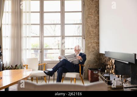 Senior man reading newspaper sitting by window in living room Stock Photo