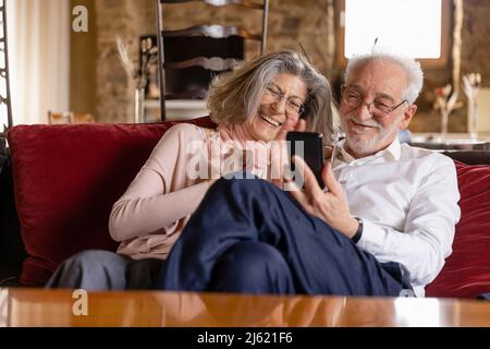 Happy senior couple having video call on mobile phone sitting on sofa at boutique hotel Stock Photo