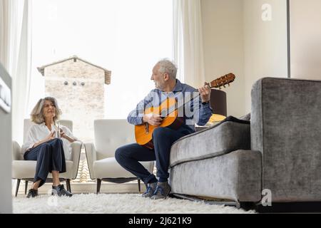 Senior woman looking at man playing classical guitar sitting on sofa at home Stock Photo