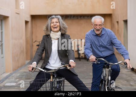 Happy senior couple riding bicycles outside building Stock Photo