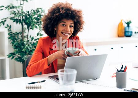 Smiling businesswoman holding coffee cup sitting with laptop at desk Stock Photo