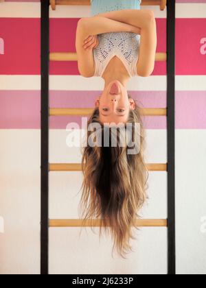 Girl sticking out tongue hanging upside down on wall bars Stock Photo
