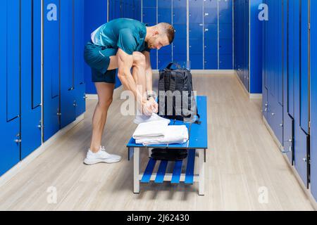 Young man tying shoe laces on bench in locker room at gym Stock Photo