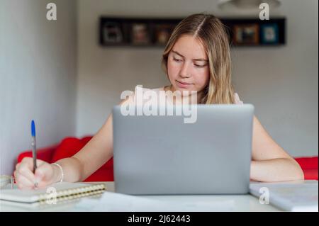 Smiling young woman writing notes sitting with laptop at home Stock Photo