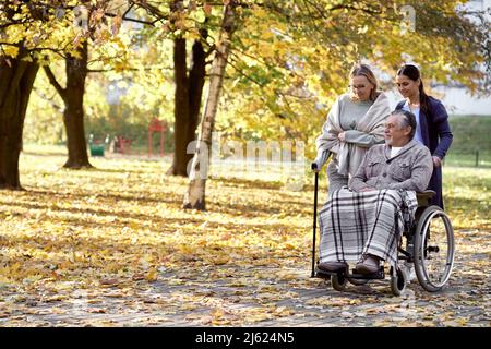 Caretaker pushing wheelchair of disabled man by woman walking with stick at park Stock Photo