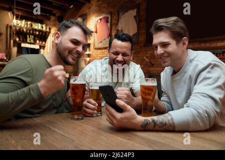 Three cheerful male soccer fans having beer and watching a match on smartphone in a pub Stock Photo