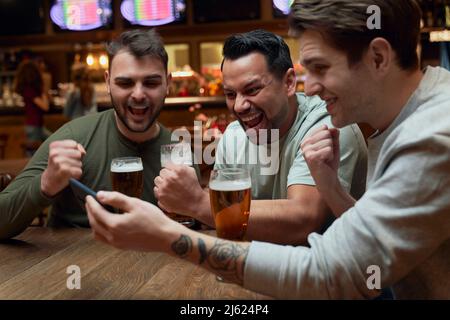 Three cheerful male soccer fans having beer and watching a match on smartphone in a pub Stock Photo