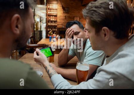 Three frustrated male soccer fans having beer and watching a match on smartphone in a pub Stock Photo