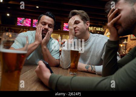 Three frustrated male soccer fans having beer and watching a match on smartphone in a pub Stock Photo