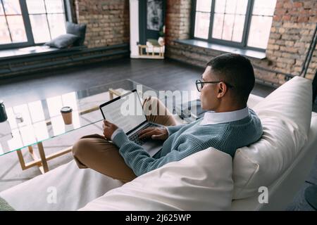 Young freelancer wearing eyeglasses using laptop in living room at home Stock Photo