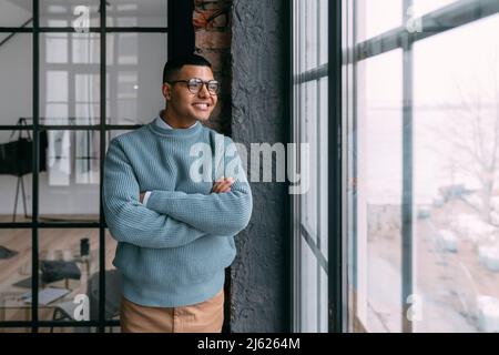 Smiling young man standing with arms crossed looking through window at home Stock Photo