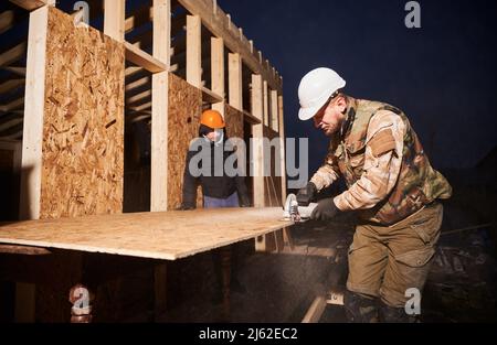 Carpenter using circular saw for cutting wooden OSB board. Man worker building wooden frame house on pile foundation. Carpentry concept. Stock Photo