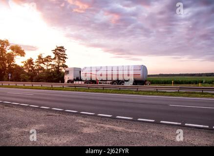 A truck with a semi-trailer tanker transports dangerous goods against the backdrop of a sunny sunset and the sky in the clouds. Transportation of flui Stock Photo