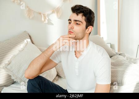 Thoughtful young man sitting on the bed in the bedroom, touching his chin and thinking. Thoughtful mature man resting on bed at home feeling sad think Stock Photo