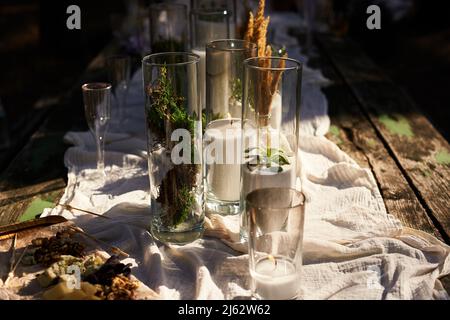 Wedding party banquet outdoors in forest. Dining table decorated in boho style with candles, white cloth, flowers, served with plates, tableware Stock Photo