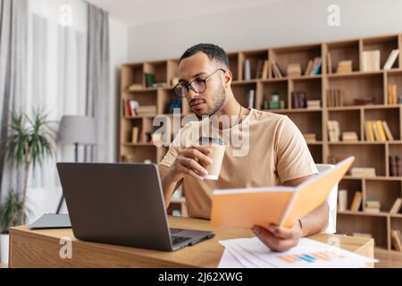 Middle eastern male student learning remotely, reading book, browsing internet on laptop and drinking coffee at home Stock Photo