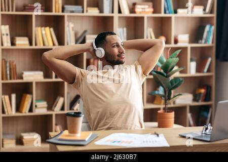 Relaxed arab man listening to music in headphones, resting with hands behind head while working on laptop at home Stock Photo