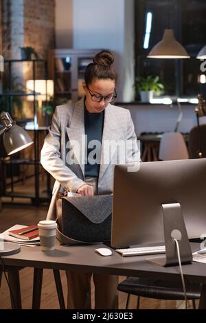 Serious young mixed race manager in glasses standing at table with computer and taking work staff while leaving office at night Stock Photo