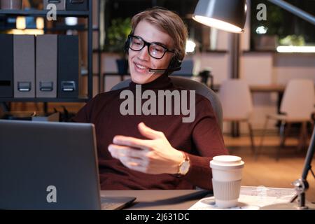 Smiling young call center operator in eyeglasses talking to customer via headset while using laptop in office at night Stock Photo