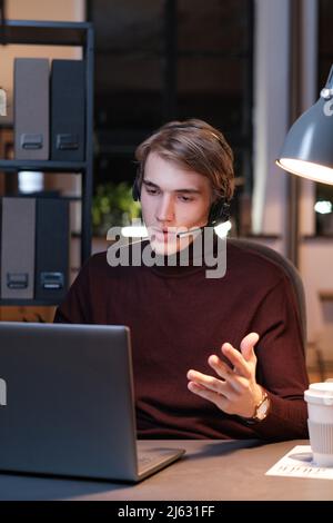 Serious young Caucasian hotline operator in headset with microphone answering online call while working with client at night Stock Photo