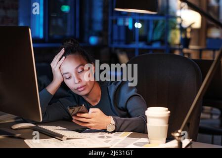 Exhausted young biracial woman sitting at table and leaning head on hand while checking phone message in office at night Stock Photo