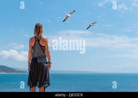 Caucasian blonde woman standing in front of the sea contemplating the horizon next to seagulls Stock Photo