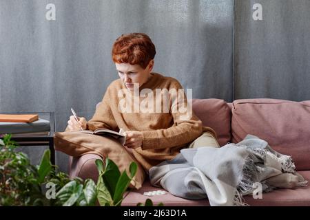 Horizontal shot of mature Caucasian woman having cold staying at home working on something making notes in notebook Stock Photo