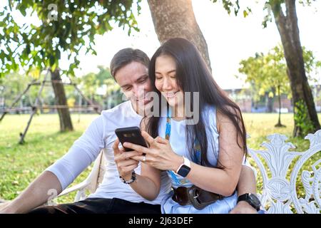 Smiling couple using the mobile while sitting on a bench outdoors during sunset Stock Photo