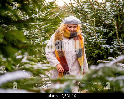 Blonde haired caucasian female wearing a beret, scarf and winter coat walking amongst snow covered Christmas trees. Stock Photo