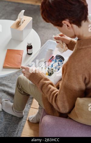 Vertical high angle shot of woman suffering influenza sitting on sofa with first aid kit on her lap looking for pills to take Stock Photo
