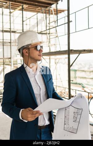 Architect wearing formal suit and hard hat holding a blueprints on a construction site Stock Photo