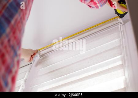 A handywoman with a tape measure installs white roller blinds indoors. Shutters day and night Stock Photo
