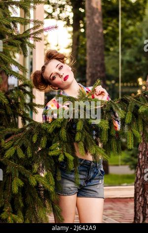 A young beautiful girl in the style of pin up stands in a park near a pine tree Stock Photo