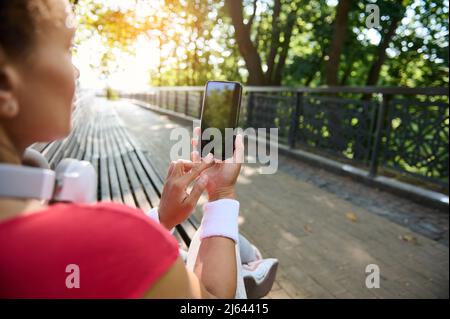 Fitness App. Sporty fit woman holding and pointing at smartphone with blank black screen while resting after training outdoors, demonstrating a copy s Stock Photo