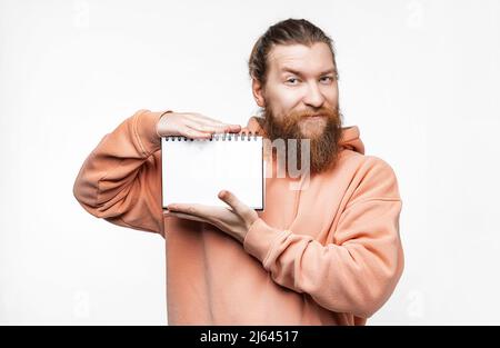 Scandinavian handsome happy man holding notepad with a white paper mockup isolated on gray background. Guy with ginger hairstyle and beard in a sweats Stock Photo