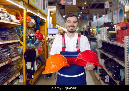 Friendly salesman demonstrates two colored safety helmets in industrial warehouse store. Stock Photo
