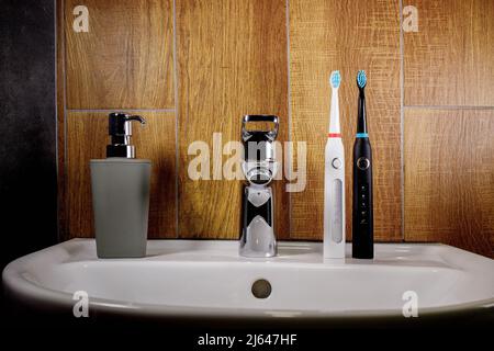 Two electric toothbrushes on the sink in the bathroom with a gray soap dispenser. Chrome-plated faucet and sink. Bathroom interior and accessories con Stock Photo
