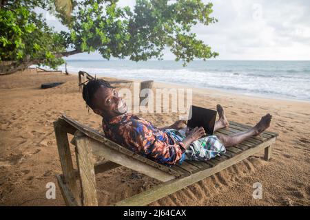 African businessman relaxes at a tropical beach on vacation while using his laptop lying on a sunbed. Stock Photo